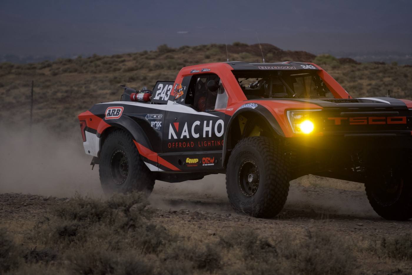An orange race truck at dusk with its headlight lit