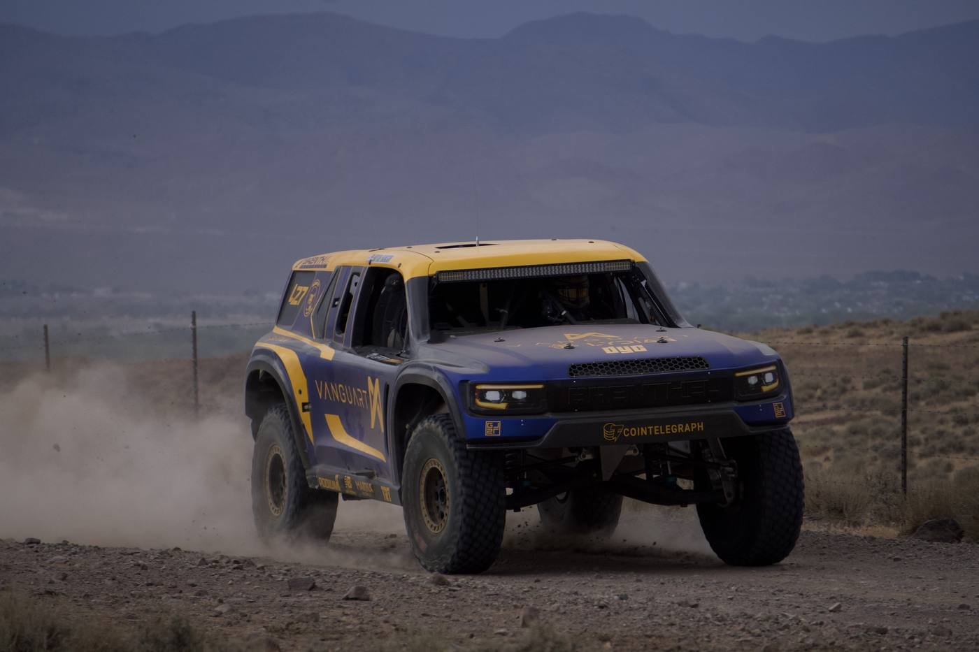 A blue and yellow race truck with a dust plume behind it