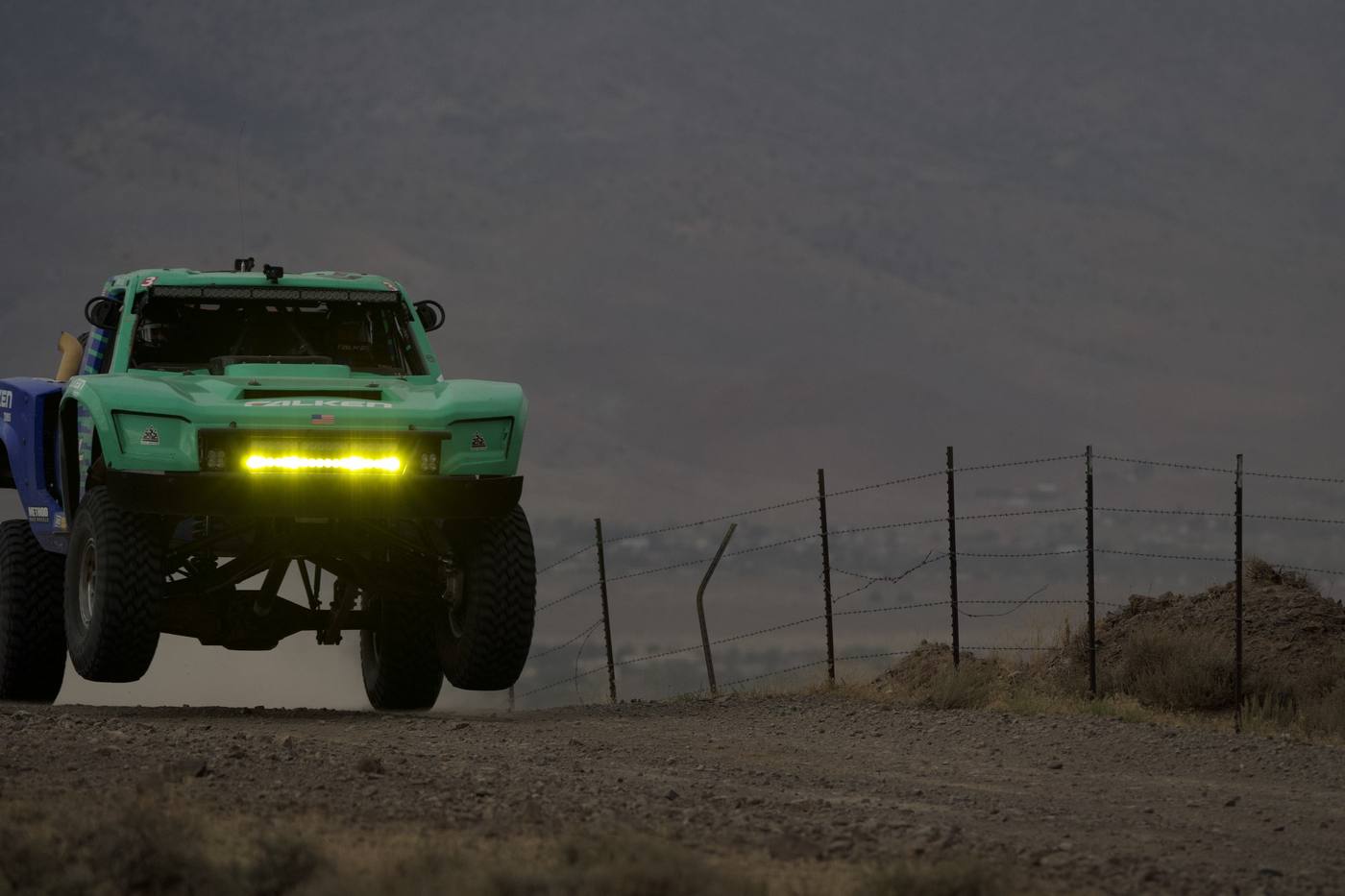 A green race truck fully airborne at dusk, light bar lit